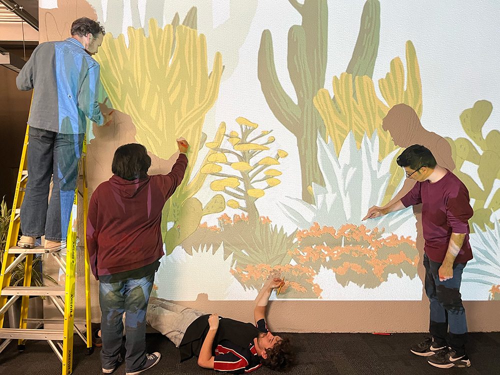 Art Club students painting a Sonoran desert mural of saguaro, agave, and prickly pear on an interior wall, working from a projected drawing